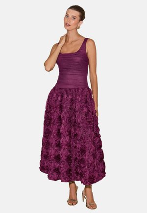 Vrouw draagt een mouwloze paarse jurk met een gestructureerde top en een volle rok met bloemmotief, gecombineerd met gouden sandalen met hoge hakken.