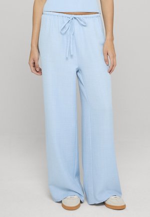 Kvinde iført lyseblå bukser med vide ben og snøre i taljen samt hvide sneakers, stående foran en ensfarvet baggrund.