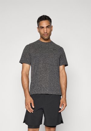 Homme se tenant devant un fond uni, portant un t-shirt de sport gris chiné à manches courtes et un short noir superposé.