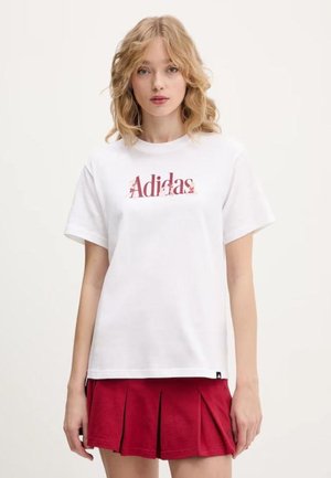 Jeune femme aux cheveux blonds portant un t-shirt blanc Adidas et une jupe plissée rouge, debout devant un fond clair uni.