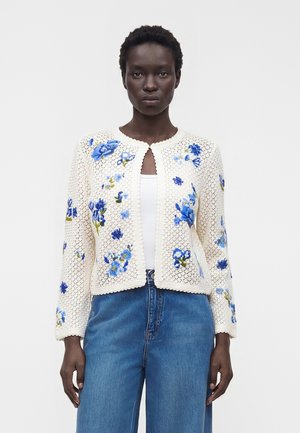 Femme portant un cardigan en crochet crème avec une broderie florale bleue sur un haut blanc et un jean bleu, debout devant un fond uni.