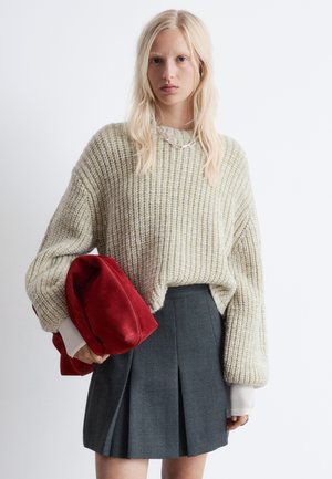 Chunky, hellbeige gestrickter Pullover mit weiten Ärmeln, kombiniert mit einem dunkelgrauen, plissierten Rock. Hält eine rot-velourene Tasche. Silberne Halsketten-Details.