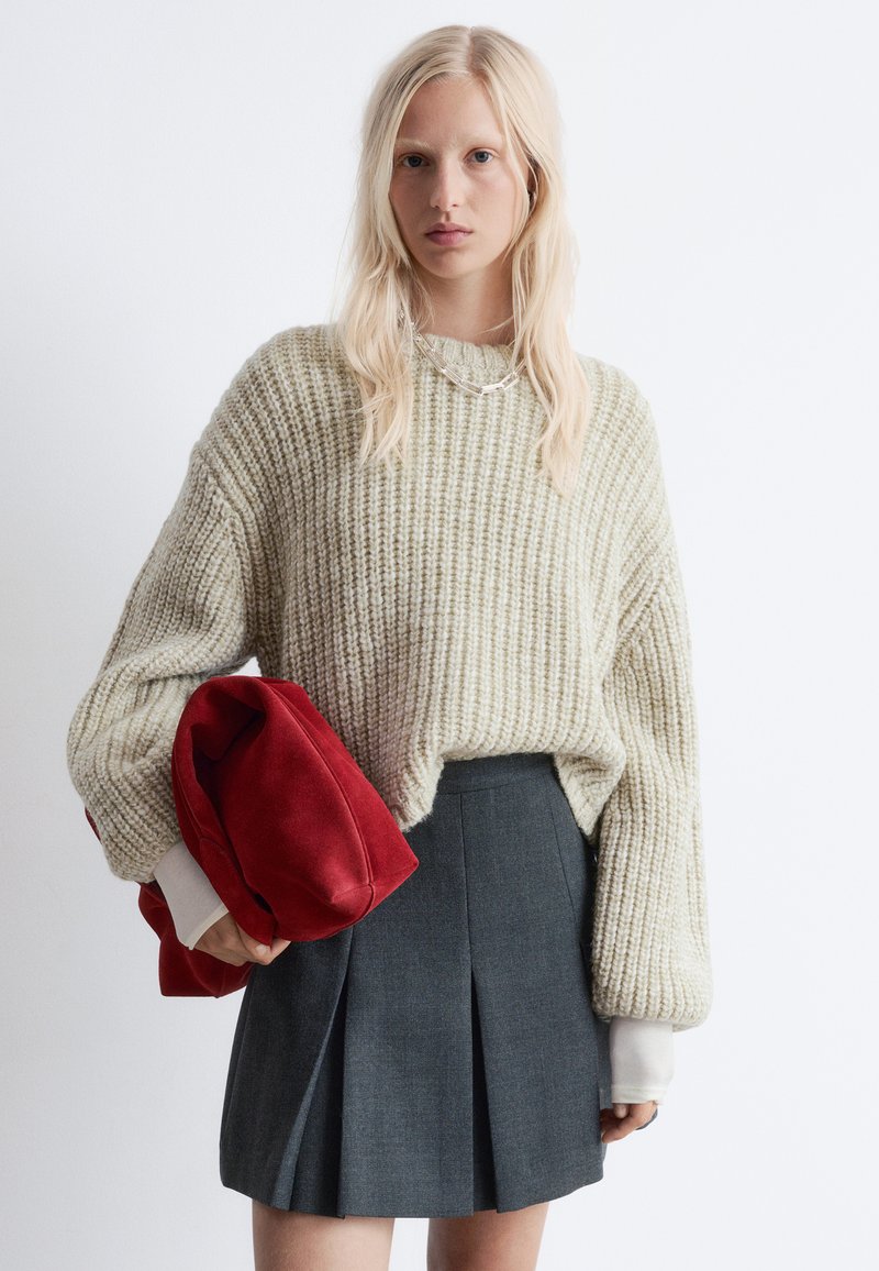 Chunky, hellbeige gestrickter Pullover mit weiten Ärmeln, kombiniert mit einem dunkelgrauen, plissierten Rock. Hält eine rot-velourene Tasche. Silberne Halsketten-Details.