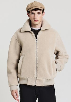 Beige Fleecejacke mit hohem Kragen, Reißverschluss vorne und gerippten Bündchen. Sie verfügt über Seitentaschen und einen lockeren Schnitt, kombiniert mit einer leichten Mütze.