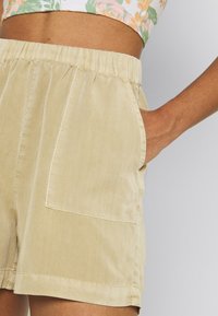 Pantaloncini beige con vita elastica, tasche laterali e una texture liscia. Presentano una vestibilità comoda e un dettaglio di orlo cucito.