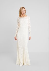 Häistä pitkähihainen mekko ivory-värisestä pitsistä, jossa on kukkakuosisia halkioita, istuva vartalo-osa ja levenevä helma. Mekossa on pyöreä kaula-aukko ja läpikuultavat hihat.