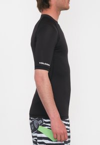 Musta lyhythihainen rash guard, jossa on istuva muotoilu ja vasemmassa hihassa valkoinen "Volcom"-logo. Yhdistetty viivallisisiin uimapöksyihin.