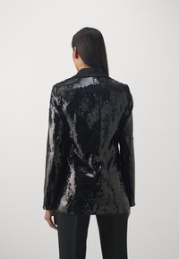 Blazer negro con lentejuelas, con un solapa negra suave, diseño ajustado y mangas largas, que exhibe una textura de superficie reflectante.