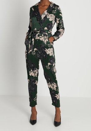 Grøn blomsterprintet langærmet slå-om-jumpsuit med midjebånd, sidelommer og sorte spidse hæle.