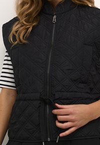 Schwarze gesteppte Weste mit hohem Kragen, vorderem Reißverschluss und Kordelzug in der Taille. Verfügt über eine kleine Brusttasche und eine Rautenmuster-Oberfläche.