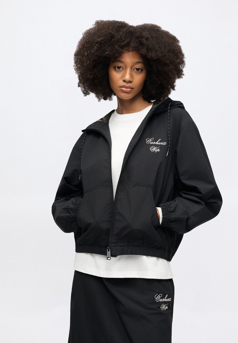 Black windbreaker jacket with elastic cuffs, zip closure, and hood. Features logo embroidery on chest and a lightweight, smooth fabric.