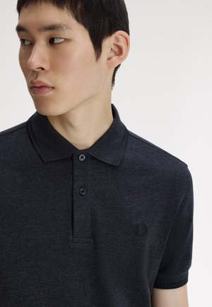 Joven girando a la izquierda, vistiendo una camiseta polo de color carbón oscuro con cuello y dos botones, que presenta un pequeño logo bordado en el pecho.