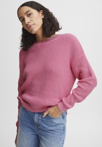Rosa Strickpullover mit lockerer Passform, Rundhalsausschnitt und gerippten Bündchen. Getragen über hellblauen Jeans, die den strukturierten Stoff und den lässigen Stil zur Schau stellen.