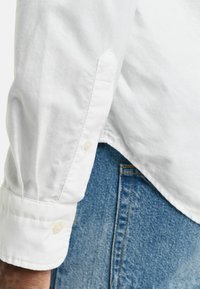 Weißes Langarmhemd mit Knopfmanschetten, das eine glatte Textur und einen leichten Glanz aufweist, kombiniert mit hellblauen Jeans.