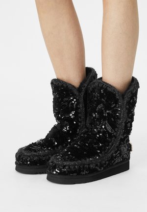 Black sequined boots with a plush, textured lining and stitched detailing; rounded top, flat sole, and a small decorative accent on the side.