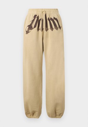 Mand iført sorte solbriller, mørkebrun jakke med lynlås, hvid tanktop, beige bukser med store sorte bogstaver og sorte sneakers, stående med hænderne i lommerne.