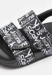 Černé sandály s bílým textem a grafickým designem "Calvin Klein Jeans". Mají nastavitelné suché zipy a strukturovanou gumovou podrážku.