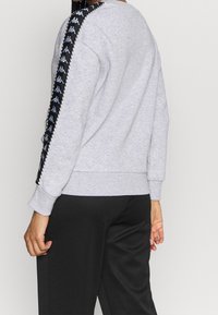 Persona vistiendo un sudadero gris claro con franjas de logo negras en las mangas y pantalones negros, de pie contra un fondo liso.