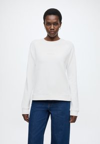 Fehér pulóver hosszú ujjakkal és bordázott mandzsettával; kerek nyakkivágással és diszkrét logóval a mellrészen. Kék farmernadrággal párosítva.