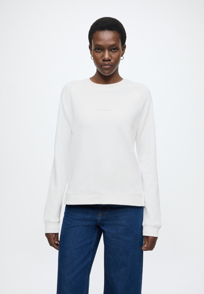 Fehér pulóver hosszú ujjakkal és bordázott mandzsettával; kerek nyakkivágással és diszkrét logóval a mellrészen. Kék farmernadrággal párosítva.