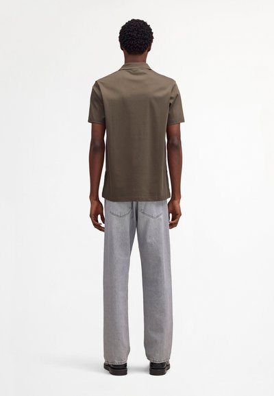 Homme aux cheveux courts et bouclés portant une chemise à manches courtes vert olive, un jean gris clair à jambes larges et des chaussures noires, debout de dos.