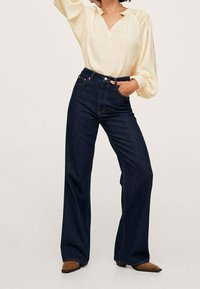 Blusa leggera color crema con maniche a sbuffo e scollatura a V, abbinata a jeans a gamba larga di colore blu scuro e stivali alla caviglia con punta affusolata marroni.