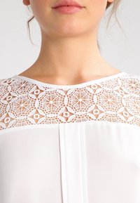 Blouse blanche avec un empiècement en dentelle à motifs géométriques et un col rond, complétée par un tissu lisse et léger.
