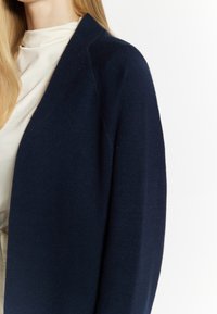 Donkerblauwe cardigan van een zachte wolmix, met een ontspannen pasvorm en een open voorkant, met minimale naden, die een lichtgekleurde top eronder aanvult.
