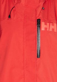 Giacca impermeabile rossa con una grande tasca con zip nera e un logo stampato di grandi dimensioni. Realizzata in tessuto liscio e resistente, con lacci regolabili.