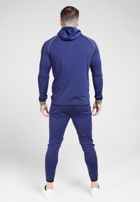 Navy hoodie and pants set made of smooth synthetic fabric, featuring a hood, fitted design, and contrasting gold accents on seams.