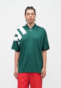 Žmogus, turintis pynimus plaukus, vilki žalią Adidas marškinėlį su baltomis juostelėmis ir raudonus šortus, priešais baltą foną.