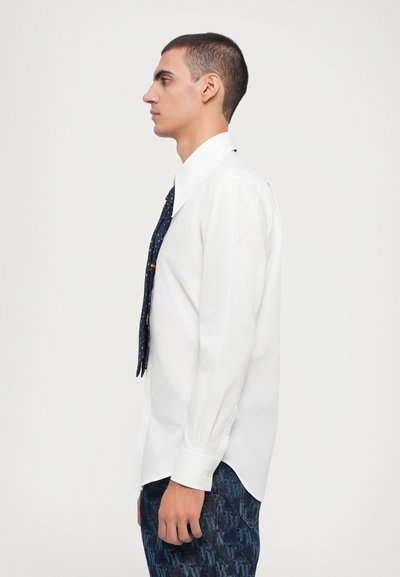 Camisa blanca de manga larga con un gran cuello, combinada con una corbata de patrón oscuro y jeans azules con patrón, vista de perfil.