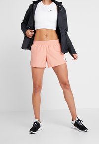 Person wearing white crop top, black jacket, peach athletic shorts, and black-and-white Nike sneakers standing against a plain background.
