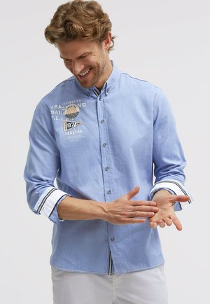 Man smiling while rolling up sleeves of light blue button-up shirt with patches and white pants, against plain background.