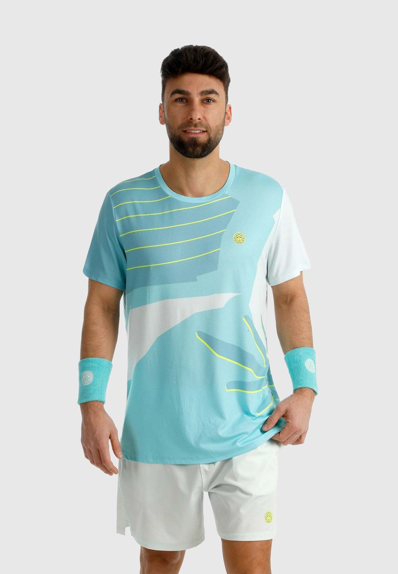 Homme portant une tenue de sport bleu clair et blanche avec des bracelets, debout devant un fond uni, face à l'objectif avec un léger sourire.