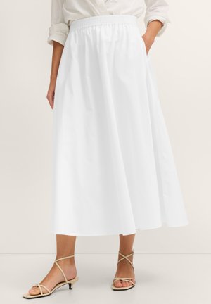 Frau trägt weißen elastischen Midi-Rock und beige Riemchensandalen mit niedrigem Absatz, steht mit einer Hand in der Tasche.