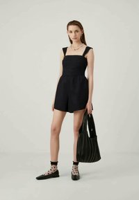 Rompers noirs avec décolleté carré et bretelles épaisses, présentant un corsage ajusté et un short ample. Le modèle tient un sac fourre-tout à rayures noires.
