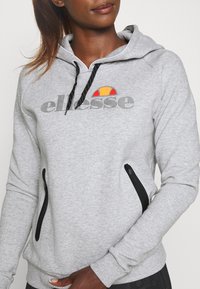 Donna che indossa un hoodie grigio ellesse con lacci neri e tasche con zip, in piedi contro uno sfondo bianco semplice.