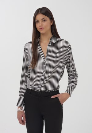 Camicetta con righe bianche e nere, con maniche lunghe e colletto, abbinata a pantaloni neri. La camicetta presenta una tasca e una texture liscia.