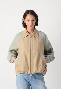 Beige bomber bunda so svetlozelenými rukávmi, so zipsom a rebrovými manžetami. Obsahuje dve predné vrecká a klasický dizajn goliera.