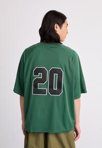 Green mesh sports jersey featuring a relaxed fit, short sleeves, and a large black number "20" at the back. Texture is breathable and lightweight.