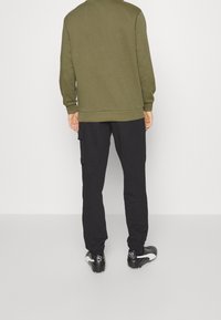 Sweat-shirt vert olive à poignets côtelés, associé à un pantalon cargo noir avec poches latérales et des chaussures de sport noires avec des accents blancs.