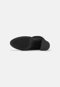 Svart mockaboot med en slät rund tå, texturerad sula och en formar häl. Har en minimalistisk design utan synlig hårdvara.