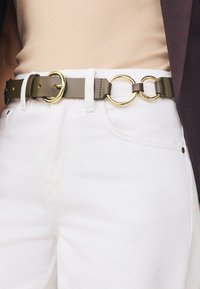 Skórzany pasek w oliwkowej zieleni ze złotymi, okrągłymi elementami ozdobnymi, z podwójnym pierścieniem, noszony na białych, dżinsowych spodniach.