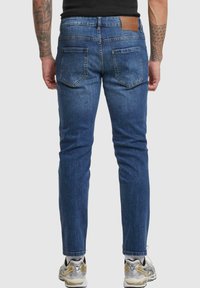 Jean en denim bleu foncé avec une coupe droite, un design à cinq poches et une étiquette en cuir au dos. Présente une finition légèrement délavée et des coutures standard.