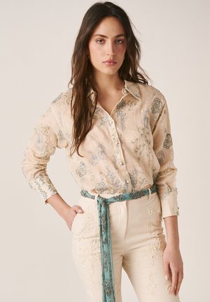 Mujer vistiendo una blusa beige con botones y estampado floral junto con pantalones crema con detalles bordados, complementados con un cinturón de tela estampada en tonos verde azulado.