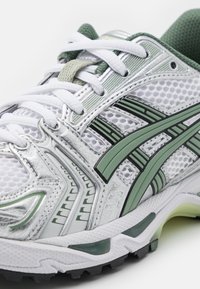 Running shoe with a white mesh upper, silver accents, green details, and black sole. Features rounded toe and contrasting striped design.