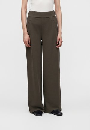 Personne portant un pantalon taille haute, jambes larges, vert olive foncé et des chaussures noires, debout devant un fond uni.