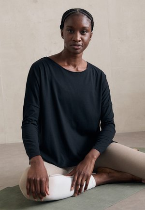Femeie cu părul împletit, așezată cu picioarele încrucișate pe un covor de yoga verde, purtând un top lung negru și colanți bej deschis, pe fundalul unei pereți simple.