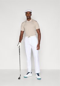 Beige polo skjorte med hvit krage, hvite bukser, hvite sko med blå detaljer og en hvit caps. Holder en golfkølle, iført hansker.